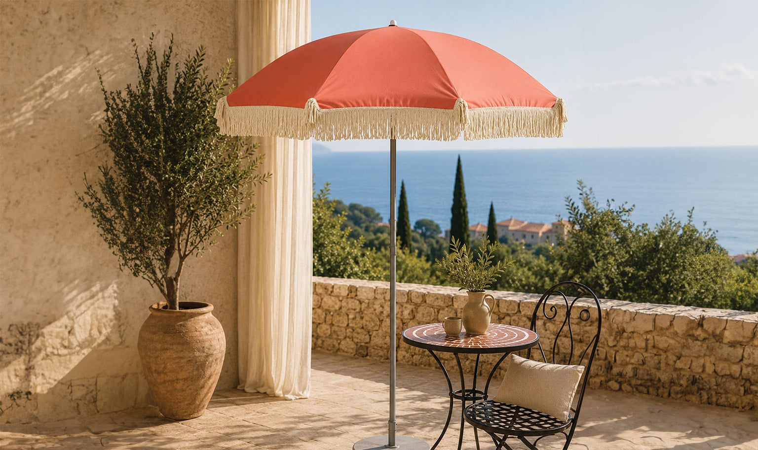 A terrace overlooking a coastal sea view featuring a coral-colored patio umbrella with decorative fringe.